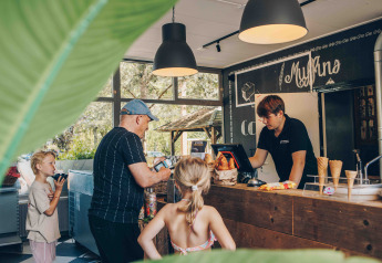Familie bestiller is og bagværk i hyggeligt glamping-kafé, med træer og varer synlige i baggrunden.