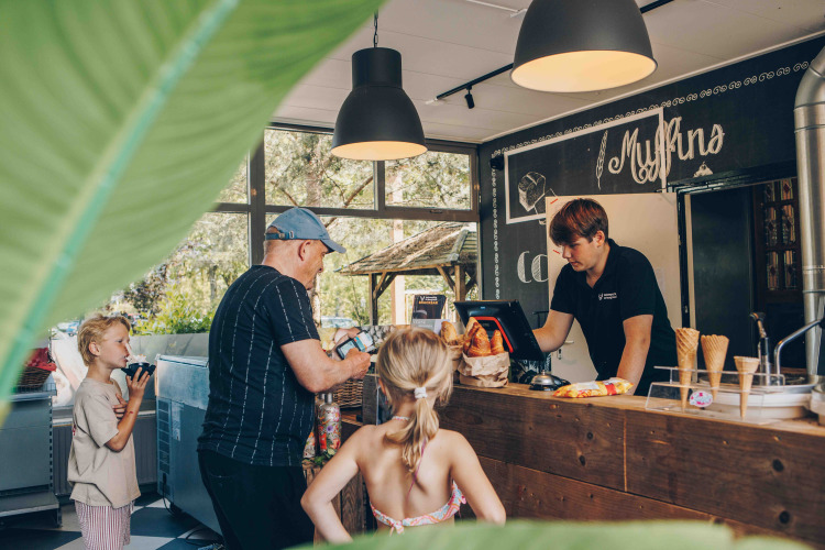 Familia compra helados y bollería en una acogedora cafetería de glamping con vegetación visible afuera.