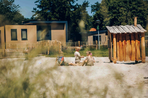 Children playing in a sandbox at a glamping site, surrounded by cabins, greenery, and wooden fences.