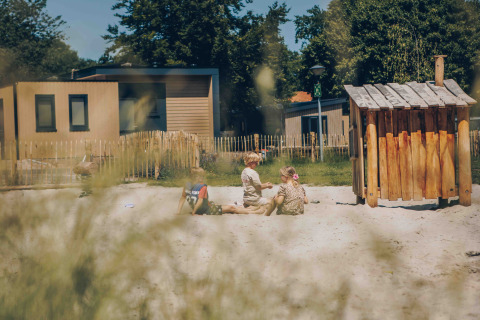 Kinder spielen im Sandkasten vor Glamping-Unterkünften, umgeben von Bäumen und Holzzäunen.