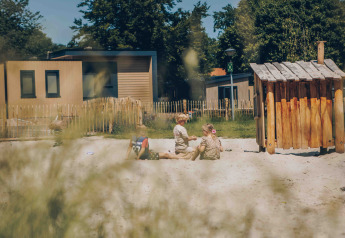 Børn leger i sandkassen ved glamping-hytter, omgivet af træer og hegn på en solrig dag.