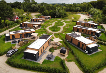 Aerial view of modern lodges surrounded by greenery with winding paths, parked cars, and forested area.