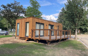 Lodge moderne en bois avec grande terrasse, entouré d’arbres et de verdure sous un ciel bleu.