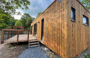 Lodge moderne en bois avec terrasse, photographiée de l’extérieur, entourée d’arbres verts par une journée ensoleillée.