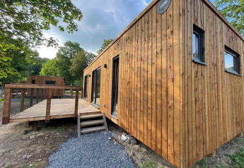 Moderne trælodge med terrasse i skovområde, set udefra på en solrig dag med grønne træer omkring.