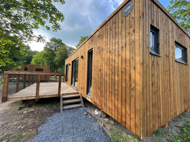 Moderne houten lodge met terras in een bosrijke omgeving, van buitenaf gefotografeerd op een zonnige dag.