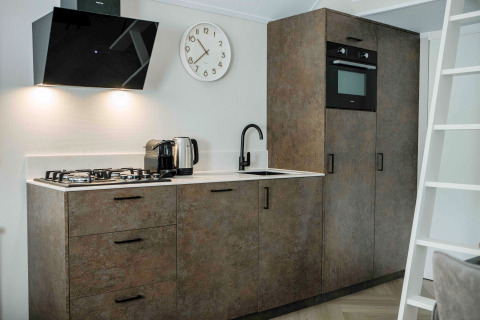 Cuisine moderne de tiny house avec cuisinière à gaz, four, bouilloire électrique, horloge et placards boisés.