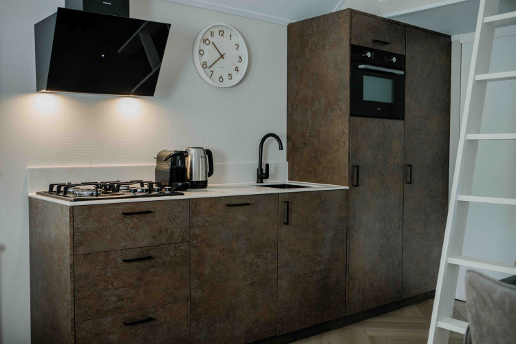 Cocina moderna en una tiny house con estufa de gas, horno, hervidor eléctrico, reloj y gabinetes rústicos.