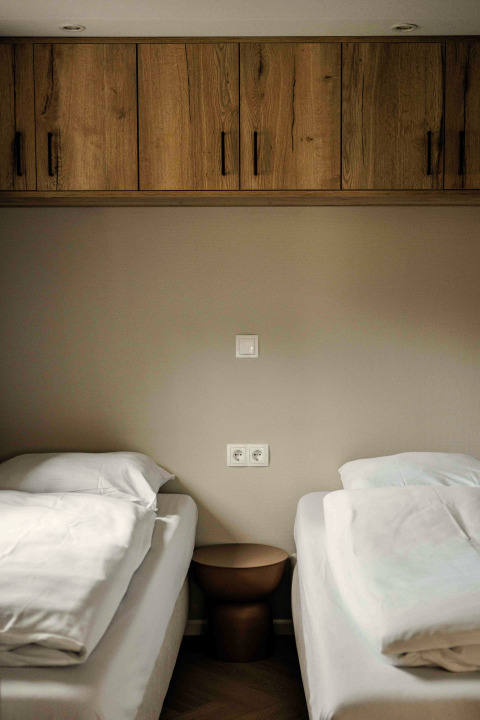 Two single beds with white bedding, a small brown nightstand and wooden cabinets in a tiny house room.