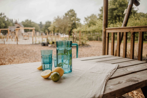 Mesa de madera al aire libre con jarra azul, vasos y limones frente a tienda safari en Veluwepark de Bosgraaf.