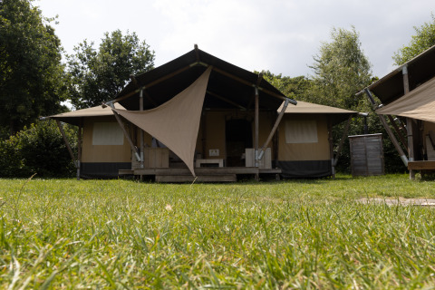 Vooraanzicht van Safari House safaritent met zonnescherm, omringd door gras en bomen en een open hemel.