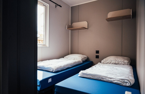 Zweibettzimmer mit blauen Matratzen, weißen Bettdecken, Wandregalen und Fenster im Loggia Wood, Eurocamping Vessem BV.
