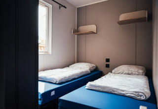 Zweibettzimmer mit blauen Matratzen, weißen Bettdecken, Wandregalen und Fenster im Loggia Wood, Eurocamping Vessem BV.