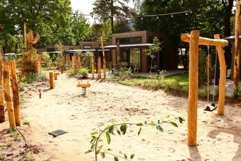 Outdoor playground with sand, swings, and wooden equipment in front of modern lodges and greenery.