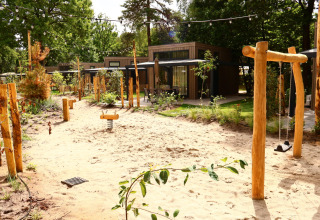Spielplatz im Freien mit Sand, Schaukeln und Holzhütten, umgeben von grüner Natur und Bäumen.