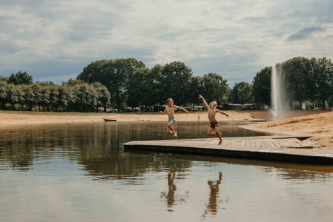 Twee kinderen springen van een houten steiger in een vijver bij een glamping met zandstrand en bomen.