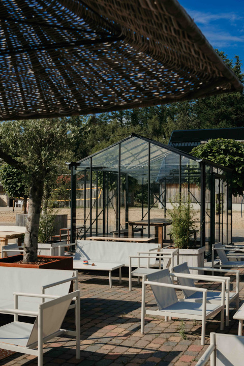 Buitenlounge van een glampingverblijf met moderne witte stoelen en een glazen serre achteraan.