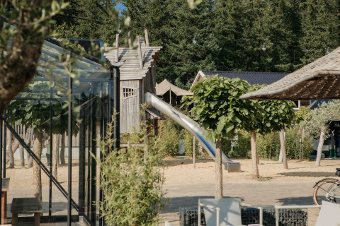 Udendørs legeplads med rutsjebane og glamping-indkvartering omgivet af træer og solrigt vejr.