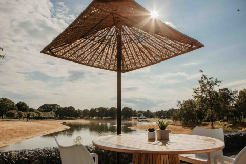 Outdoor table and chairs under a parasol with a view of the lake and trees at a sunny glamping site.