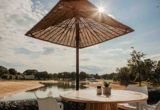 Tavolo e sedie da esterno sotto un ombrellone con vista sul lago e alberi in un glamping soleggiato.