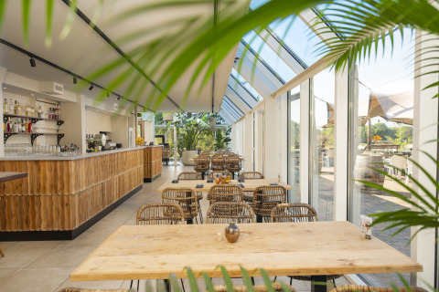 Licht en modern glampingcafé met glazen wanden, houten meubels en uitzicht op groene natuur buiten.
