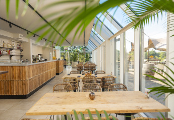 Licht en modern glampingcafé met glazen wanden, houten meubels en uitzicht op groene natuur buiten.