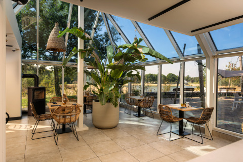 Lichte glampingaccommodatie met veel glas, tropische plant en rieten stoelen in gezellige sfeer.