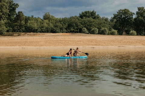 Twee mensen peddelen op een opblaasbare boot bij een zandstrand op een glampinglocatie.