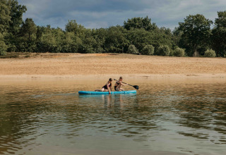 Two people paddling on an inflatable raft near a sandy beach and forest at a glamping accommodation.
