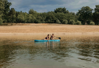Two people paddling on an inflatable raft near a sandy beach and forest at a glamping accommodation.