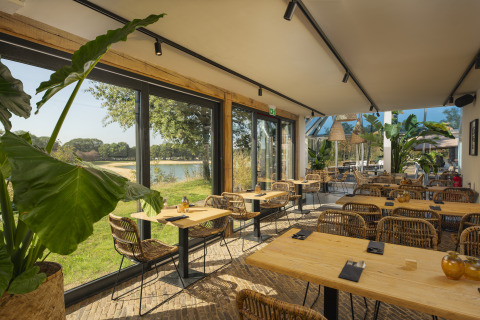 Lys restaurant med store vinduer og udsigt til natur og sø ved glamping, indrettet med kurvemøbler.