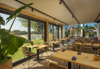Restaurant lumineux avec grandes baies vitrées et vue nature dans un site de glamping, meubles en osier.