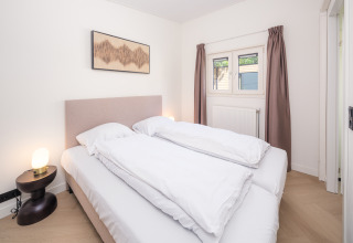 Schlafzimmer in einer Lodge mit Doppelbett, Nachttisch und Lampe, Fenster mit braunen Vorhängen.