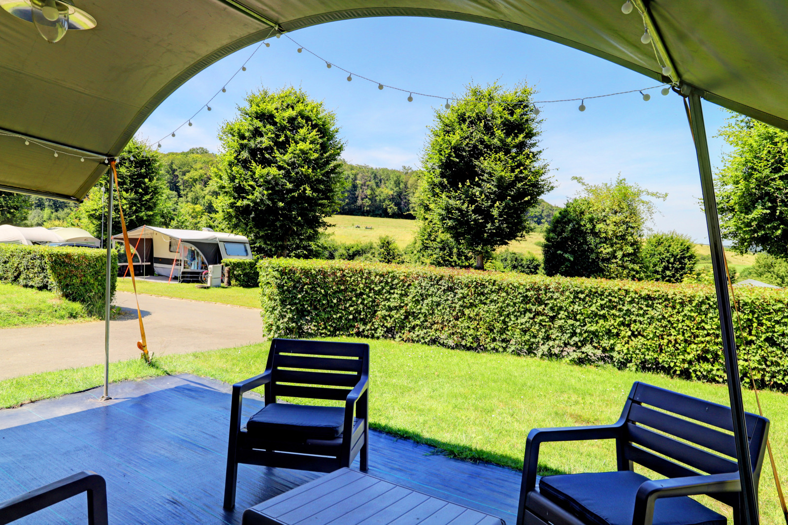 Uitzicht vanaf een lodge-terras met tuinstoelen, groene heggen en caravans op een zonnige dag in de natuur.