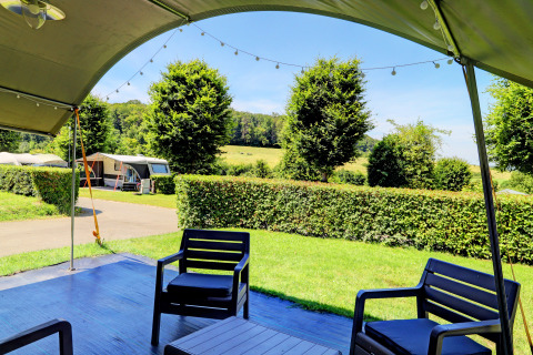 Vista dalla veranda di un lodge con sedie da giardino, siepi e roulotte su uno sfondo verde e collinare.