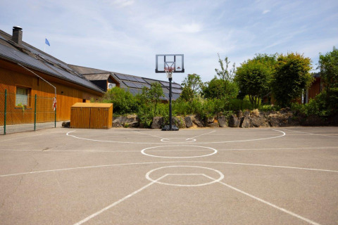 Outdoor basketball court at Camping Nommerlayen in Luxembourg, surrounded by greenery and wooden buildings.