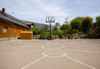 Campo da basket all'aperto al Camping Nommerlayen in Lussemburgo, circondato da vegetazione e edifici in legno.