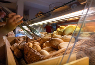 A hand uses tongs to pick freshly baked bread rolls from a basket at the bakery in Camping Nommerlayen.