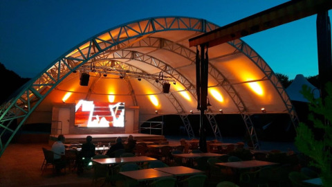 Palco all'aperto con tetto a tenda illuminato di sera al Camping Nommerlayen in Lussemburgo.