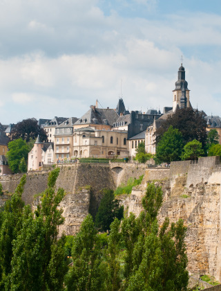 Scenic view of Luxembourg City with historic buildings and cliffs surrounded by lush green trees and sky.