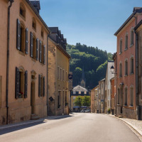 Street view in Nommern, Luxembourg, with colorful buildings and a church spire in the forested distance.