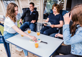 Famiglia si diverte con un gioco da tavolo e succhi all’aperto, seduta davanti a un lodge in vacanza.