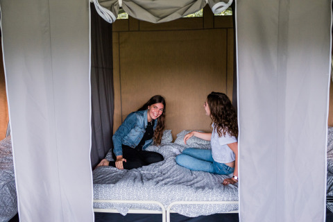 Due giovani donne sorridono e chiacchierano sedute su un letto in una lodge, circondate da tende.