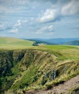 Rolling grassy hills and a steep rocky cliff near Herpelmont, Grand Est, France, under a partly cloudy sky.