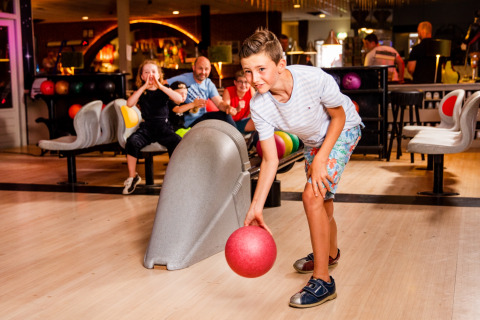 Jongen aan het bowlen in een speelhal, terwijl anderen op de achtergrond enthousiast toekijken en aanmoedigen.