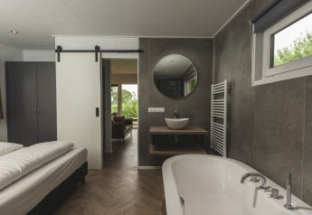 Suite moderna al River Lodge 'Wellnesshuus' presso Holiday Park Mölke con vasca, lavabo, letto e porta scorrevole.
