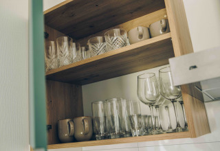 Openkast in een tiny house, toont ordelijk geordende glazen, kopjes en wijnglazen op houten legplanken.