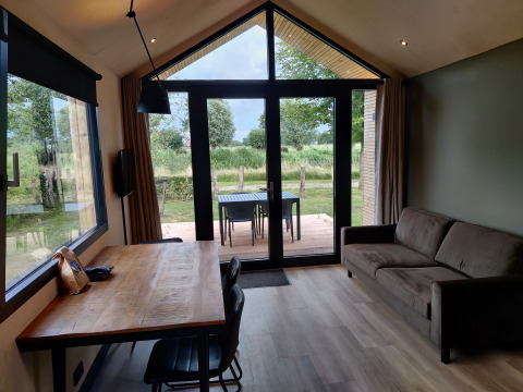 Vue intérieure d'un lodge confortable avec canapé, table à manger et grandes fenêtres sur la nature verte.
