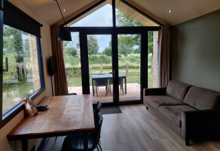 Vue intérieure d'un lodge confortable avec canapé, table à manger et grandes fenêtres sur la nature verte.