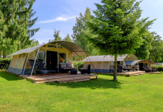 Afbeelding van moderne tenten met houten terrassen bij een groene lodge, omgeven door bomen en gras.